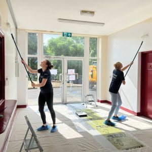 two people with long painting rollers paint the entrance hall to a building