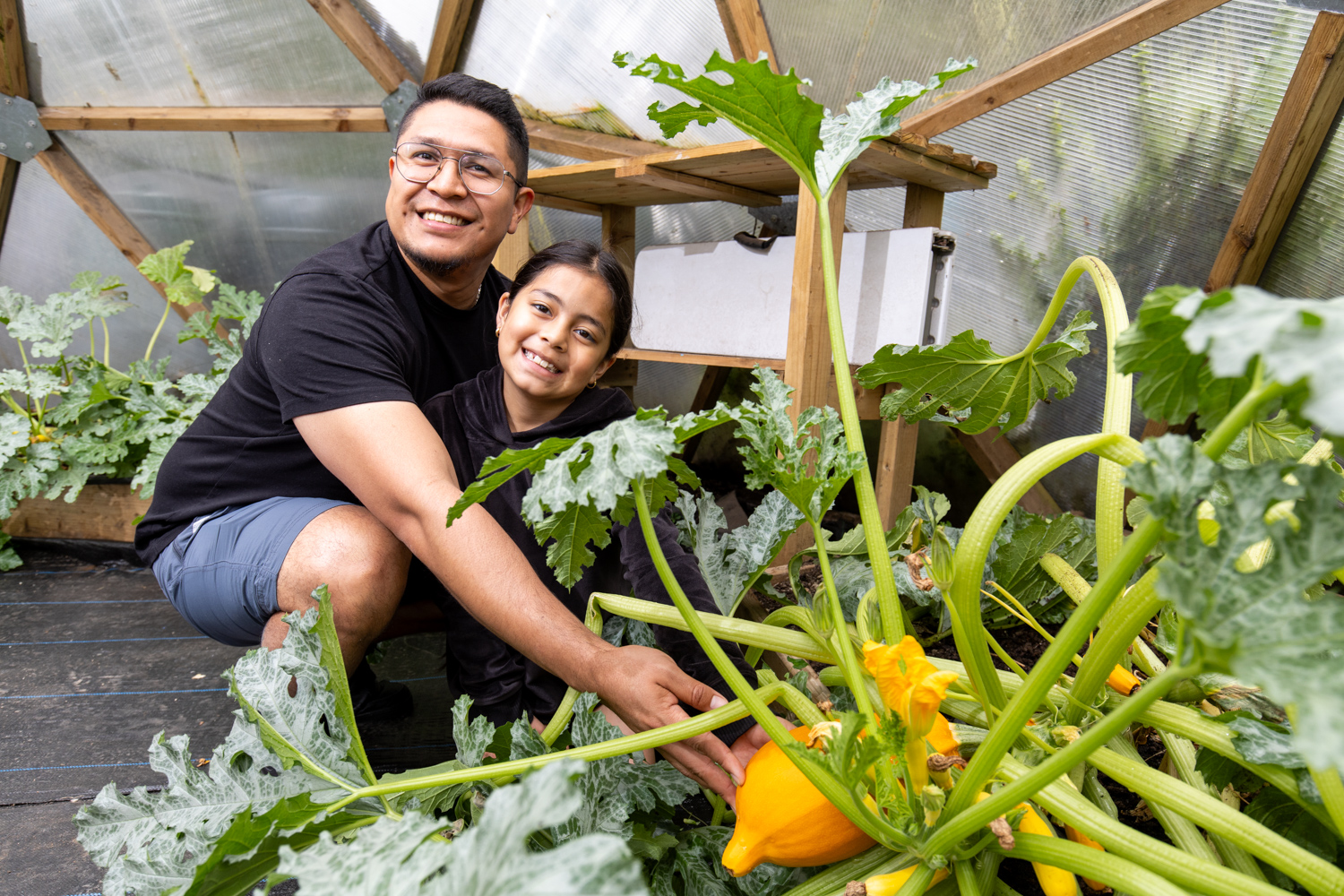 a man and his young daughter kneel by an impressively large squash plant in a garden dome