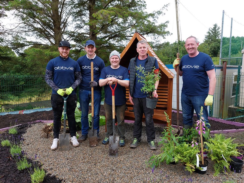 5 smiling men hold gardening tools in front of a shed